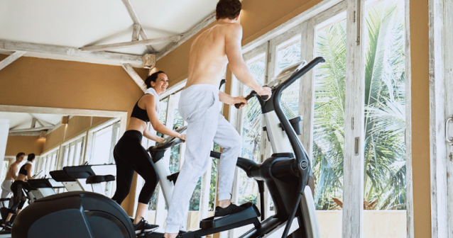 Man and woman exercising on elliptical and treadmill machines inside a bright gym with large windows and tropical palm trees outside.